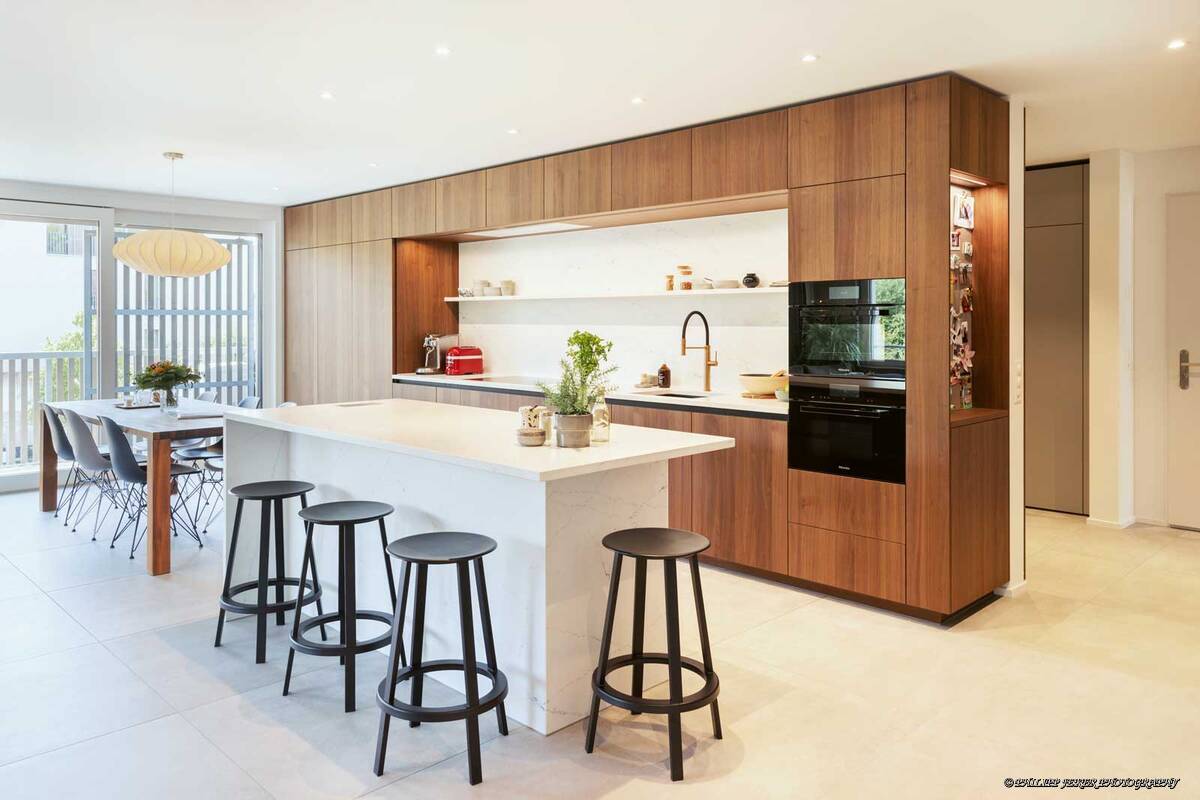 Die Kombination aus Holz und Marmor symbolisiert eine Einheit von Eleganz, Natürlichkeit und Moderne. Das Holz setzt warme Akzente und schafft eine einladende Atmosphäre, während die Kücheninsel aus Marmor durch seine glatte Oberfläche sanft und luxuriös das einfallende Licht reflektiert. Sanitas Troesch AG.